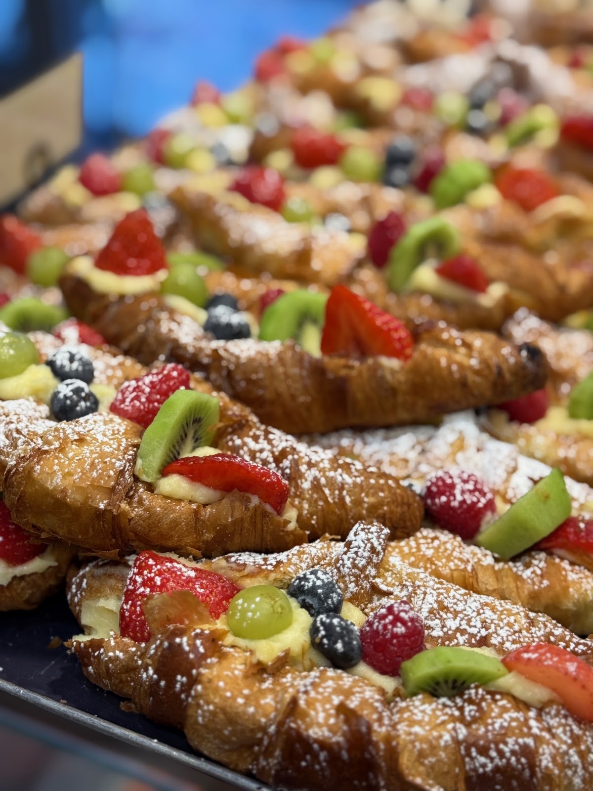 A tray of croissants filled with pastry cream and topped with strawberries, kiwi, grapes and blueberries under a heavy dusting of icing sugar.
