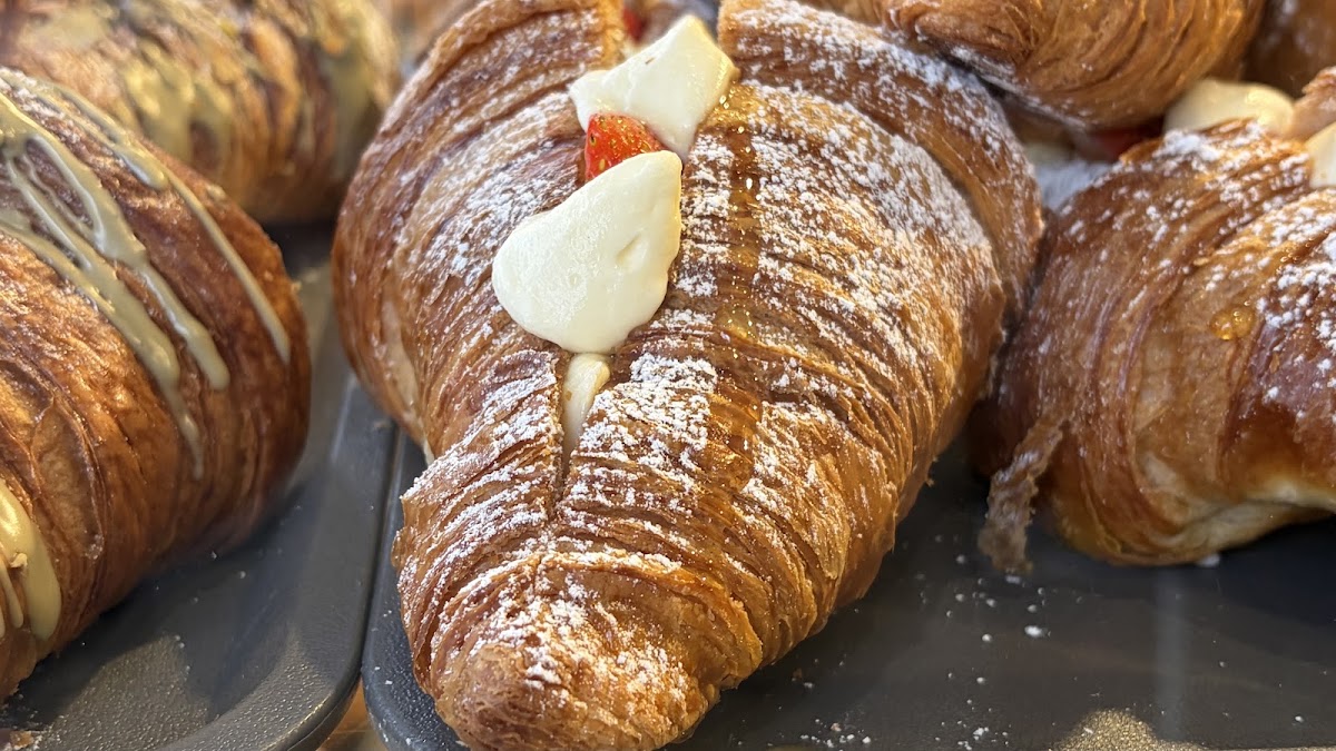 A close-up of a laminated croissant filled with cream and a slice of strawberry, dusted with icing sugar, on a dark baking tray.