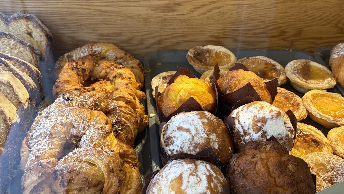 A cabinet shelf holding twisted nut-topped pastries, sliced marbled loaf, muffins in dark paper cases and a tray of custard tarts.