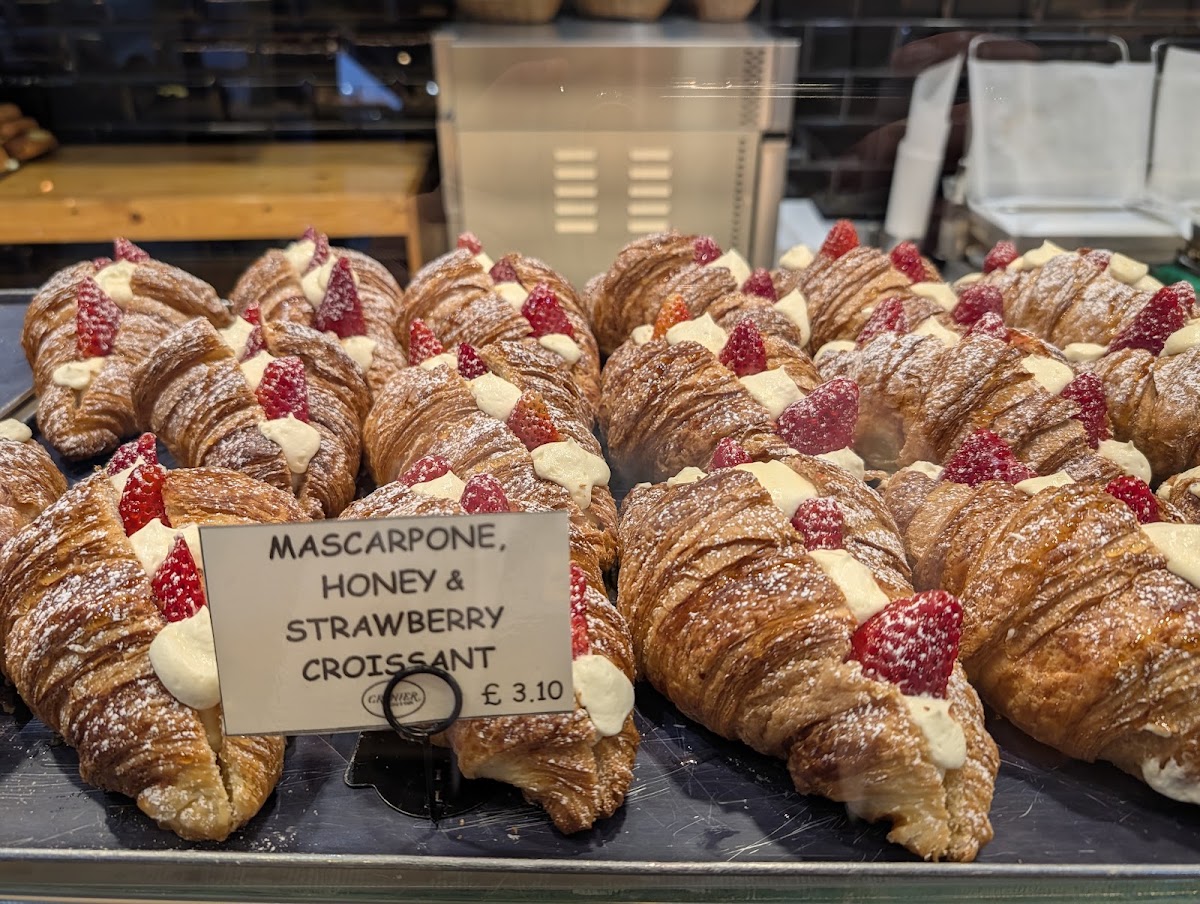 A metal tray of croissants split and filled with white cream and strawberries, dusted with icing sugar, beside a handwritten card reading Mascarpone, Honey and Strawberry Croissant.