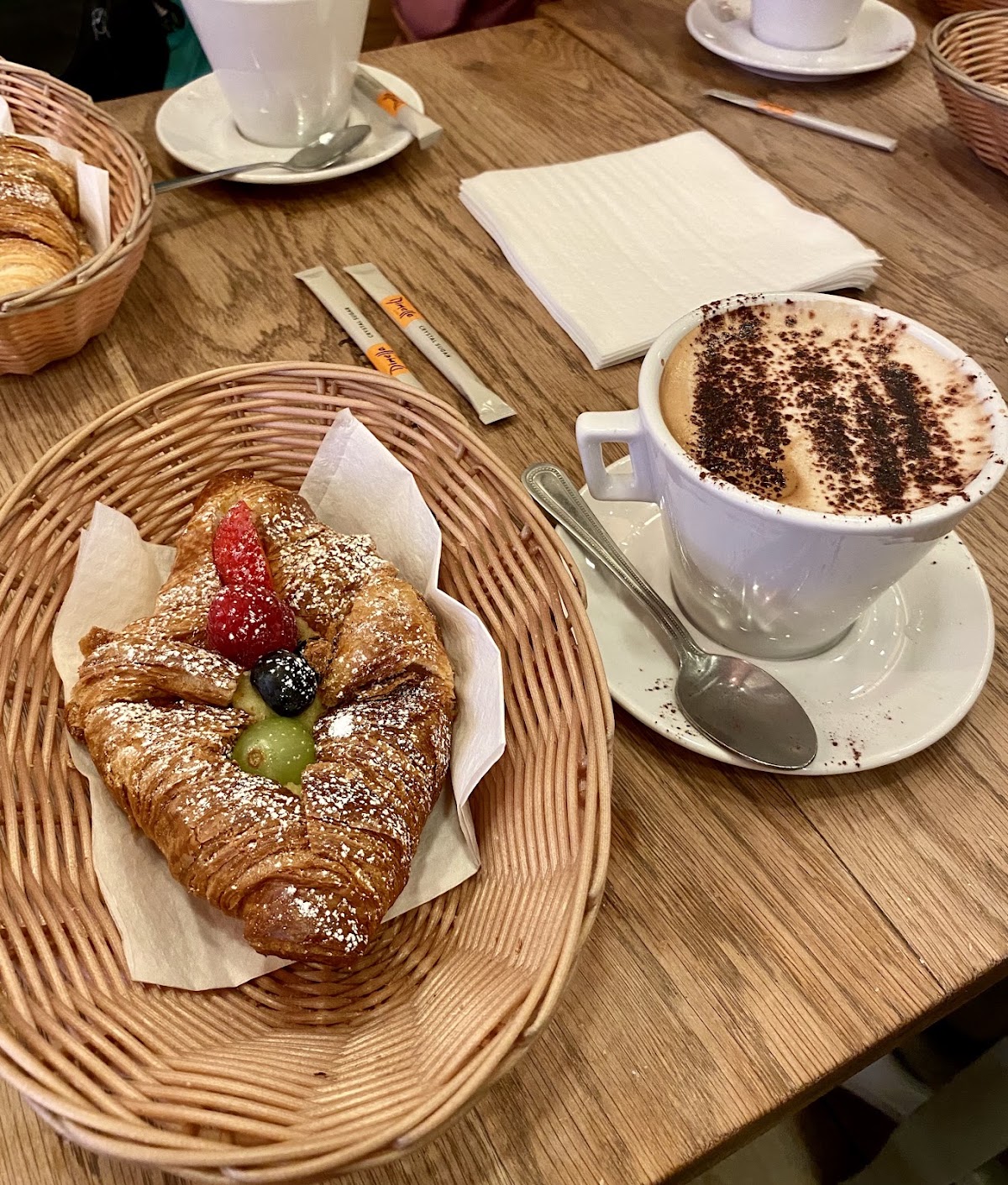 A fruit-topped croissant in a wicker basket lined with paper, next to a cappuccino dusted with cocoa in a white cup and saucer, on a wooden table.