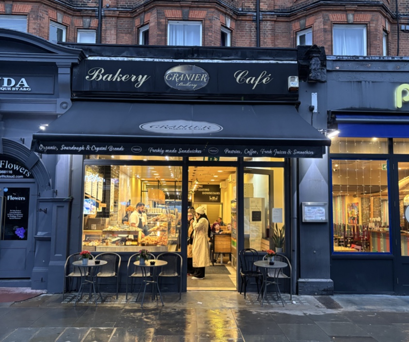 The shop front at 115 Queensway at dusk: a dark grey fascia lettered Bakery on the left and Café on the right, with an oval Granier Bakery sign between them, a lit interior with a serving counter, and small pavement tables under the awning.